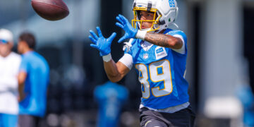 Football player in a blue jersey (#89) reaching to catch a spiraling ball, white helmet with yellow facemask in action.