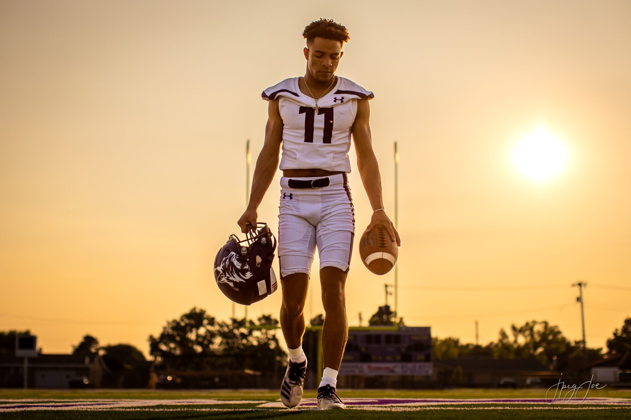 brenen-02 | Press Pass Sports Football player in a white jersey number 11 walks on a sunlit football field, carrying his helmet in one hand and a football in the other.