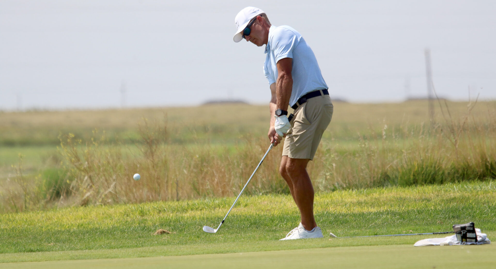 Golf35 | Press Pass Sports Golfer in light blue polo and beige shorts putting on a green fairway, preparing to strike the ball with a putter.