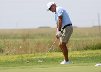 Golf35 | Press Pass Sports Golfer in light blue polo and beige shorts putting on a green fairway, preparing to strike the ball with a putter.