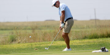 Golfer in light blue polo and beige shorts putting on a green fairway, preparing to strike the ball with a putter.