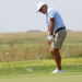 Golf35 | Press Pass Sports Golfer in light blue polo and beige shorts putting on a green fairway, preparing to strike the ball with a putter.