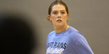 Woman with light brown hair tied back, wearing a blue athletic shirt with 'West Plains' text, looking off to her right in a gym or indoor setting.