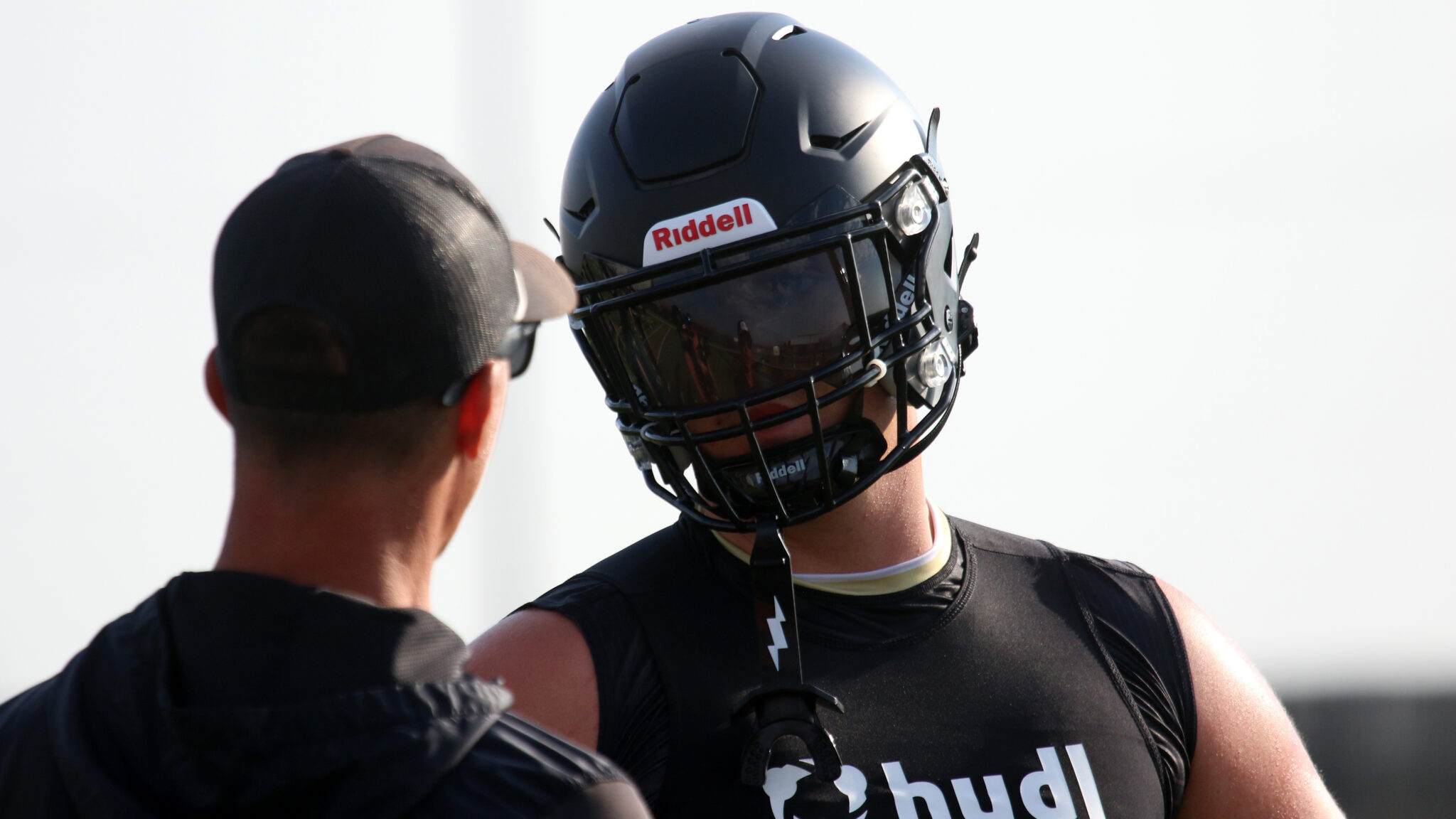 1K6A7973 | Press Pass Sports Two football players in conversation, one wearing a black Riddell helmet with dark visor and a black jersey on a bright day.
