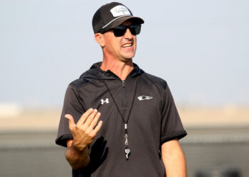 1K6A8179 | Press Pass Sports Man wearing a black athletic shirt, cap, and sunglasses speaks outdoors, gesturing with his left hand.