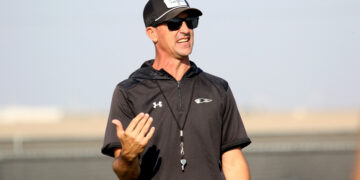 Man wearing a black athletic shirt, cap, and sunglasses speaks outdoors, gesturing with his left hand.
