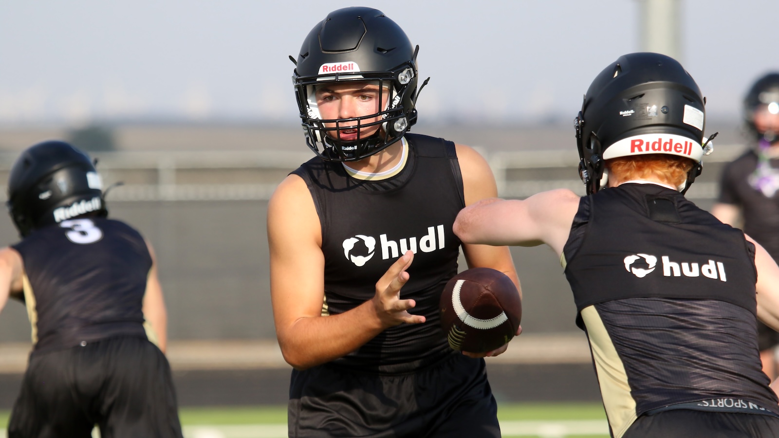 Football players in black Hudl jerseys practice on a field; central player holds a football, ready to pass, wearing a Riddell helmet.