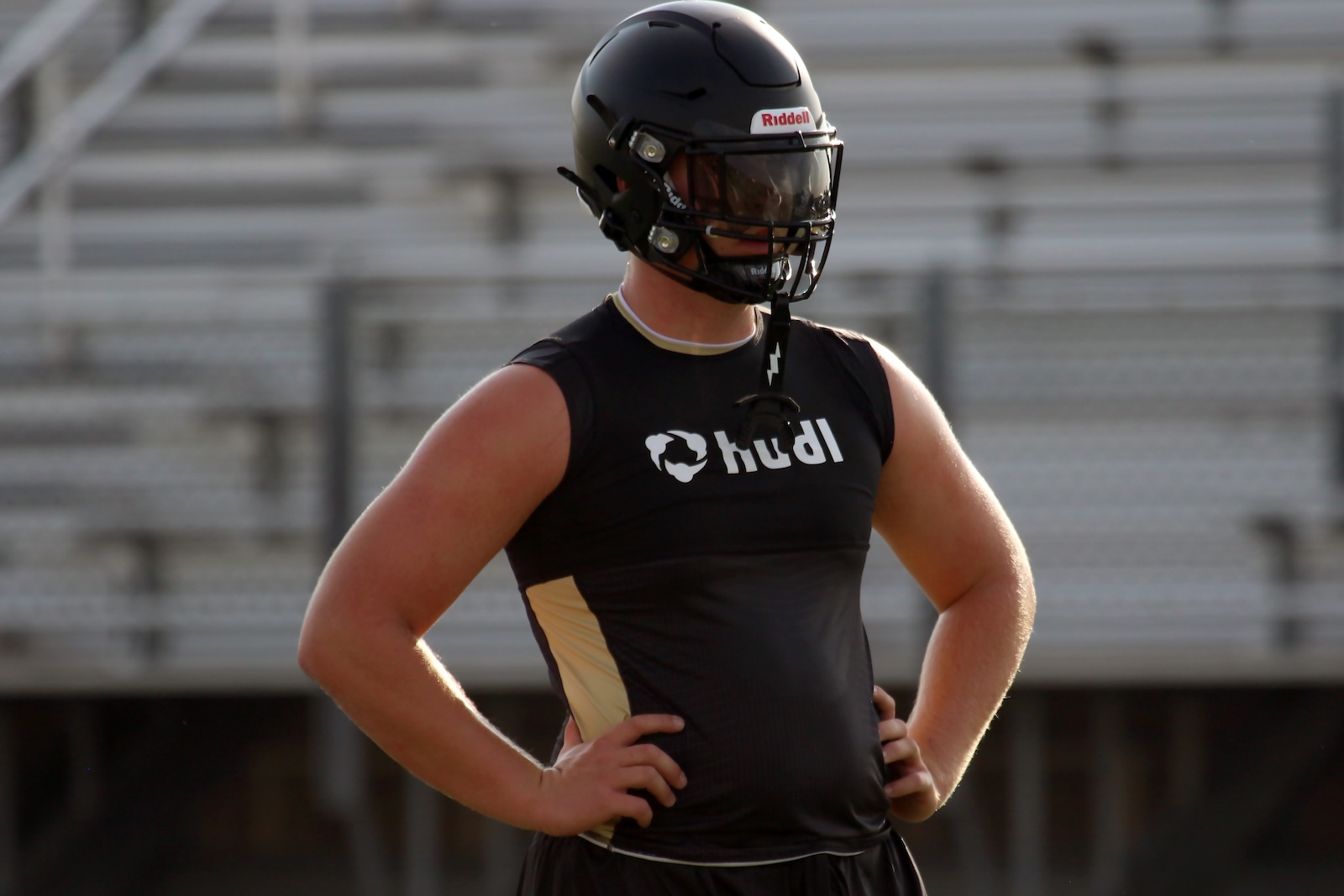 Football player in a black sleeveless jersey and black helmet, hands on hips on a bleacher backdrop.