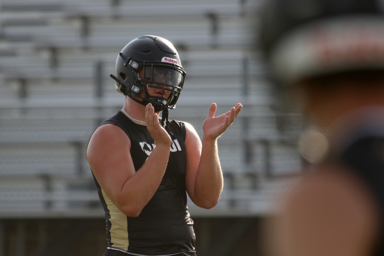 American football player in a black and gold uniform raises hands in a clapping gesture on a practice field, wearing a dark helmet with a face mask.