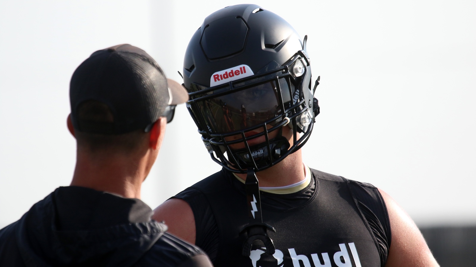 Football player in black gear wearing a Riddell helmet with visor, talking to a coach nearby.