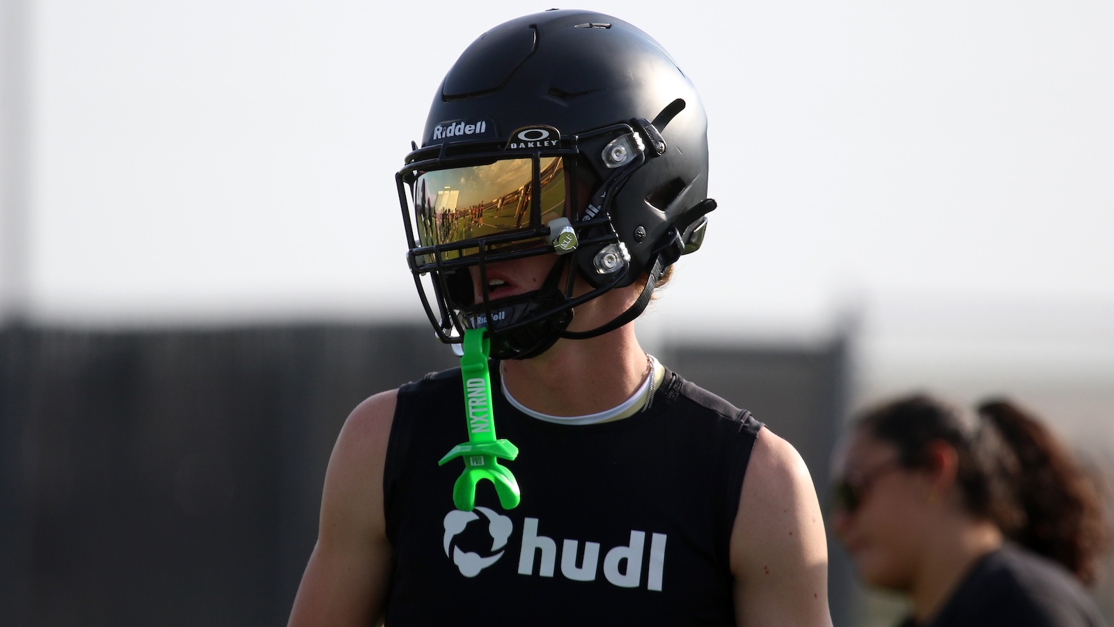 Football player in a dark helmet with a reflective visor, wearing a Hudl-branded sleeveless shirt and a bright green whistle lanyard.