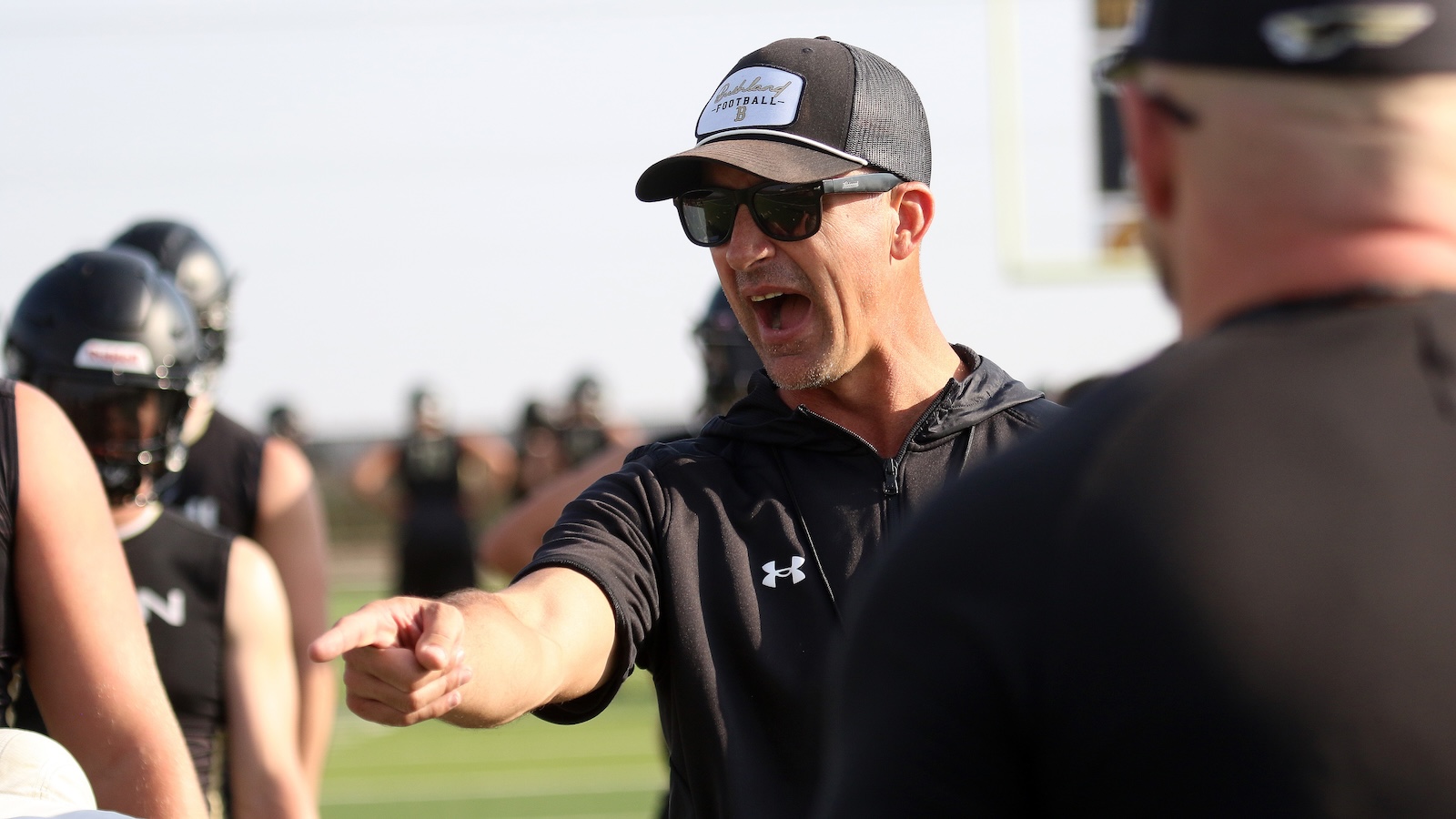 Coach shouting and pointing at players during football practice on a sunny field, wearing a black cap, sunglasses, and a black Under Armour jacket.