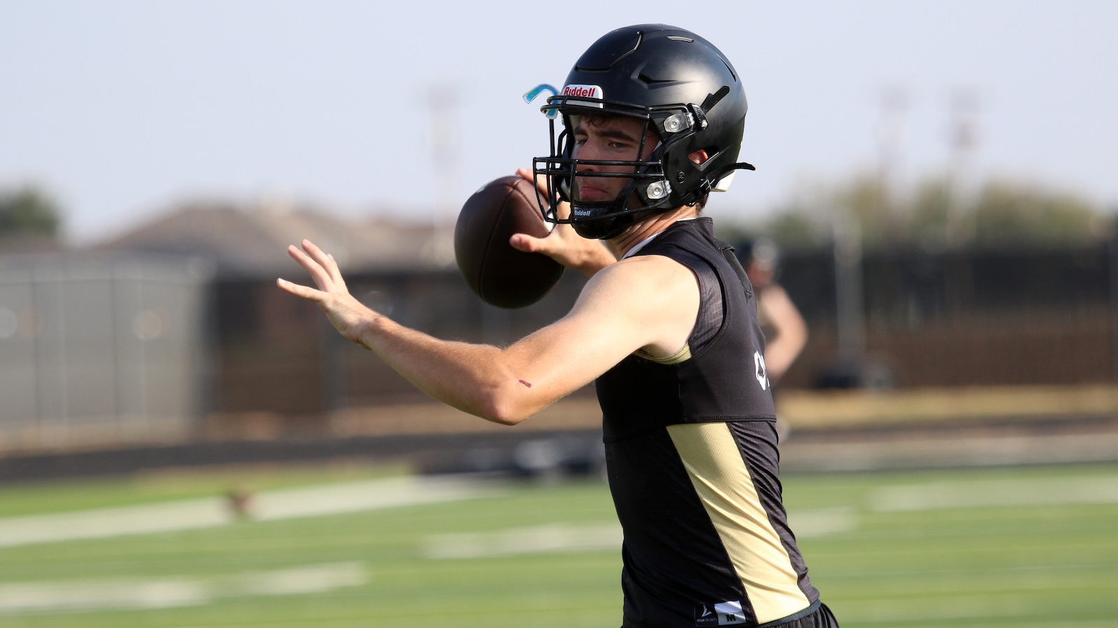 Male football quarterback in a black and gold jersey throws a football on a practice field, wearing a black helmet with a facemask.