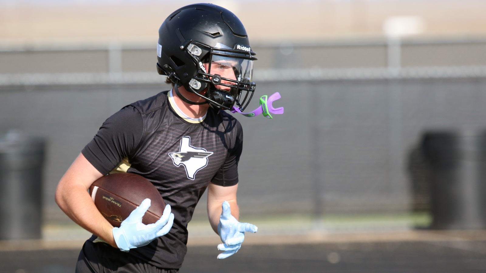 Football player in black uniform runs with ball, wearing a black helmet and light-blue gloves on a track/field.