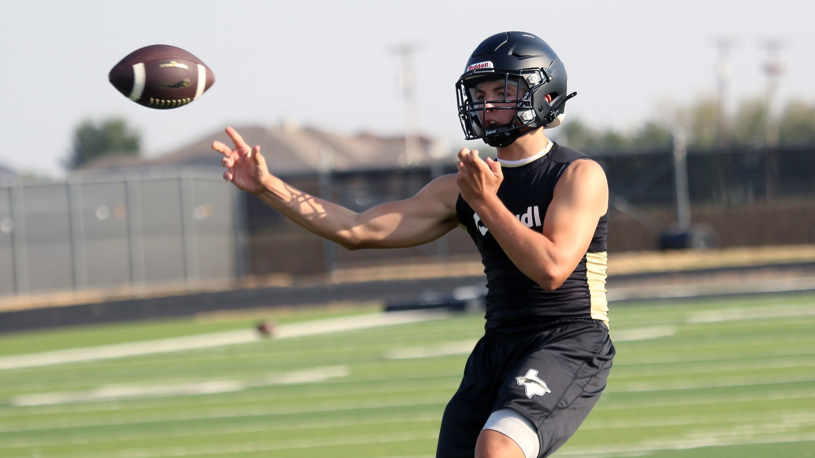 Football player in a black uniform and helmet reaches to catch a spiraling ball on a sunny practice field.