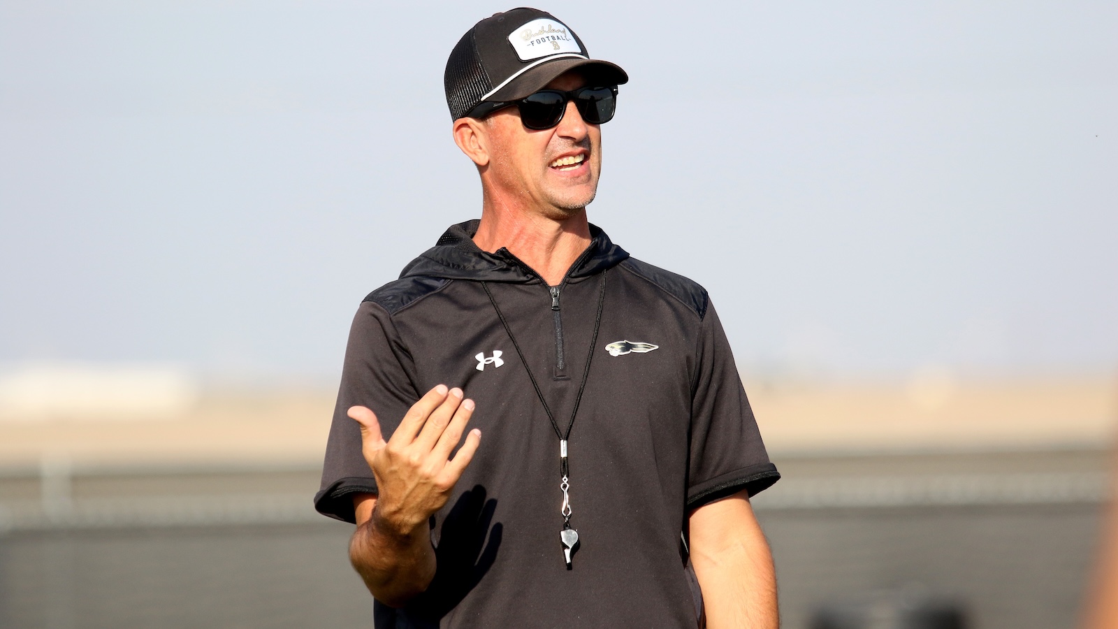 Athletic coach in a black shirt and cap, wearing sunglasses and a whistle, gesturing during practice on a sunny day.