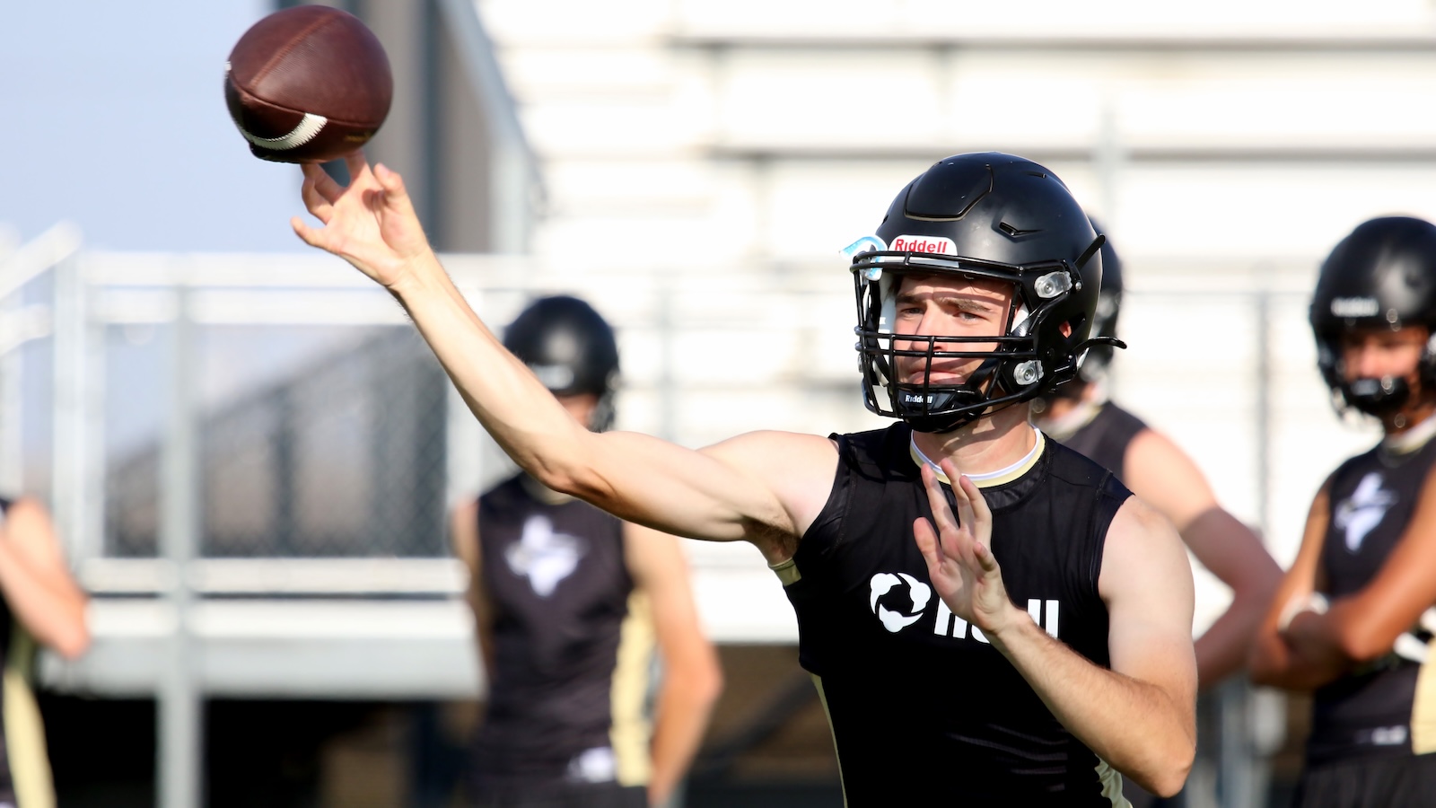 Male football player in a black practice jersey and helmet throwing a football during drills on an outdoor field.