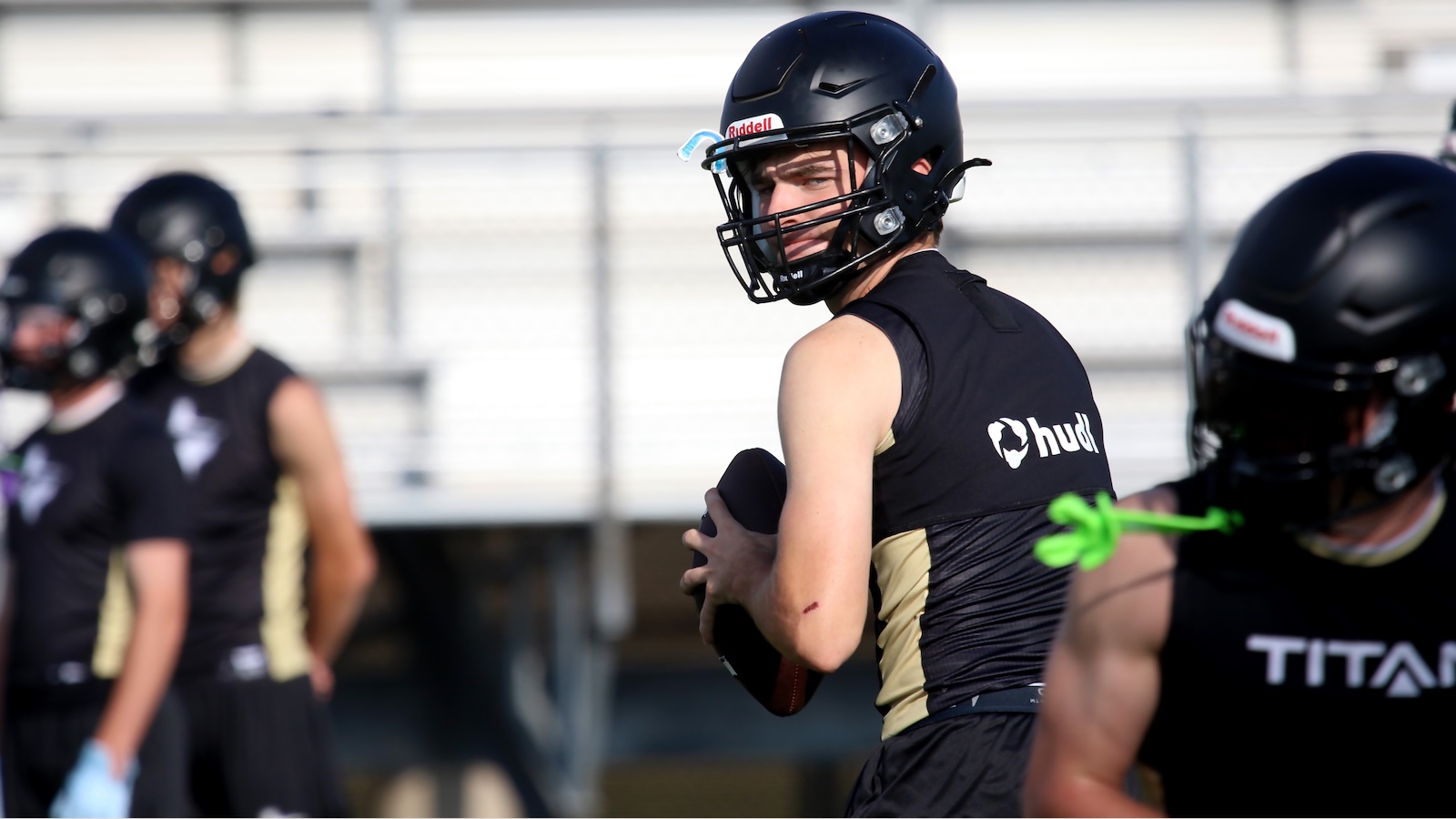Football player wearing a black helmet and jersey holds a football, looking back over his shoulder on a practice field with blurred teammates in the background.