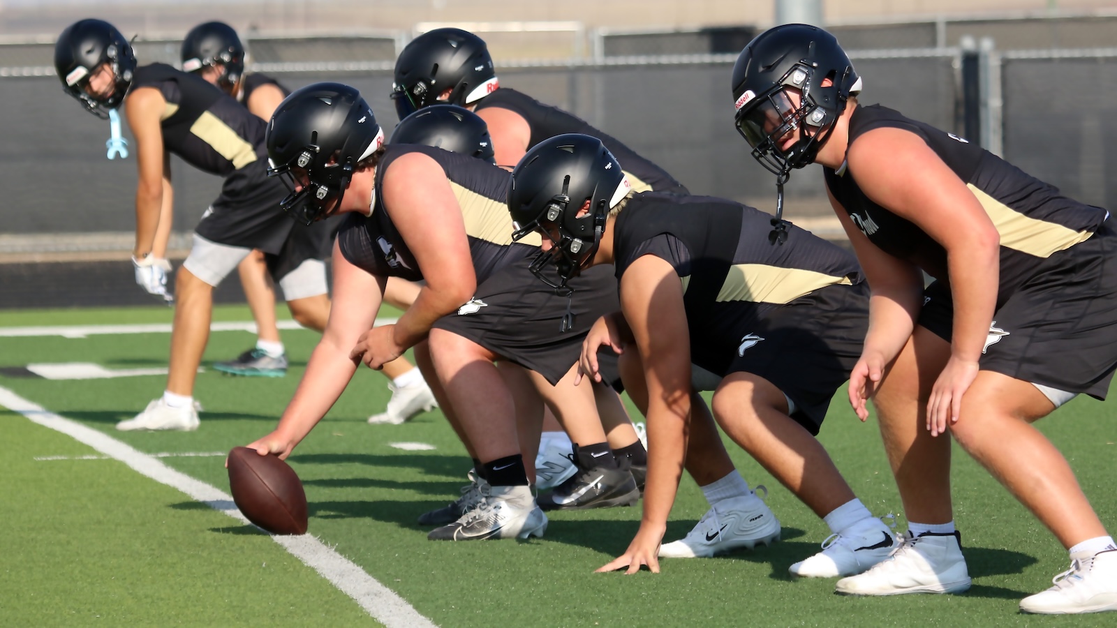 American football linemen in black and gold uniforms crouch at the line of scrimmage as the ball is snapped at practice on a green field.