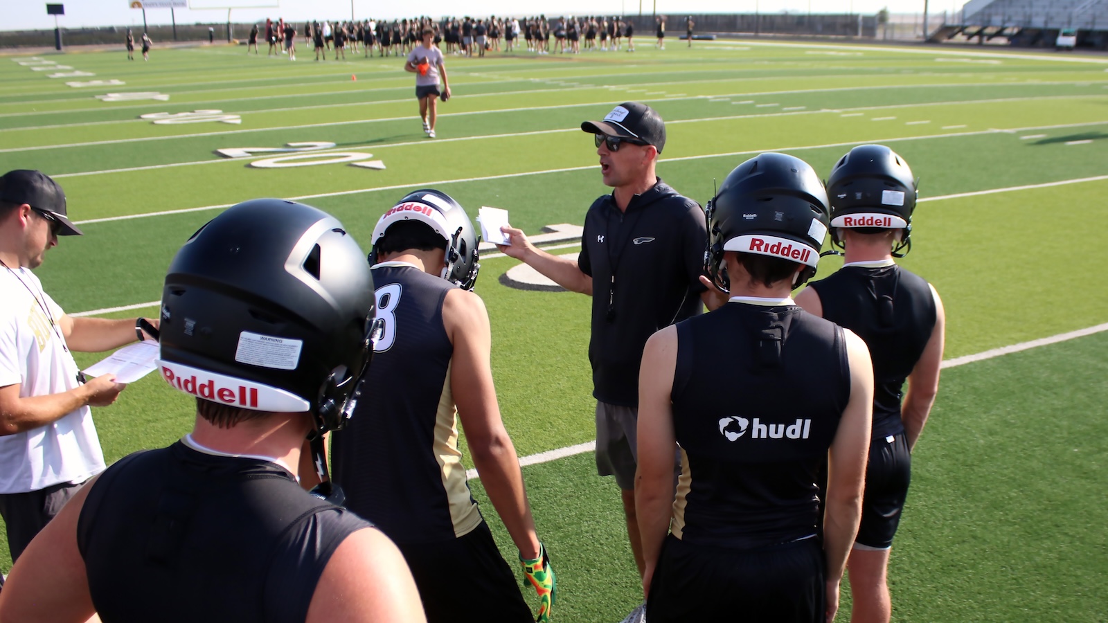Football coach talks to a group of football players in black uniforms during a practice on a sunny green field, helmets labeled Riddell and hudl logo visible.