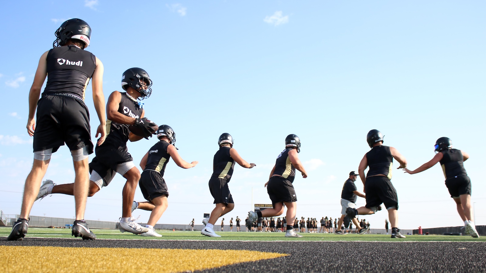 American football players in black uniforms sprint on a sunny outdoor field during practice, seen from behind with helmets on.
