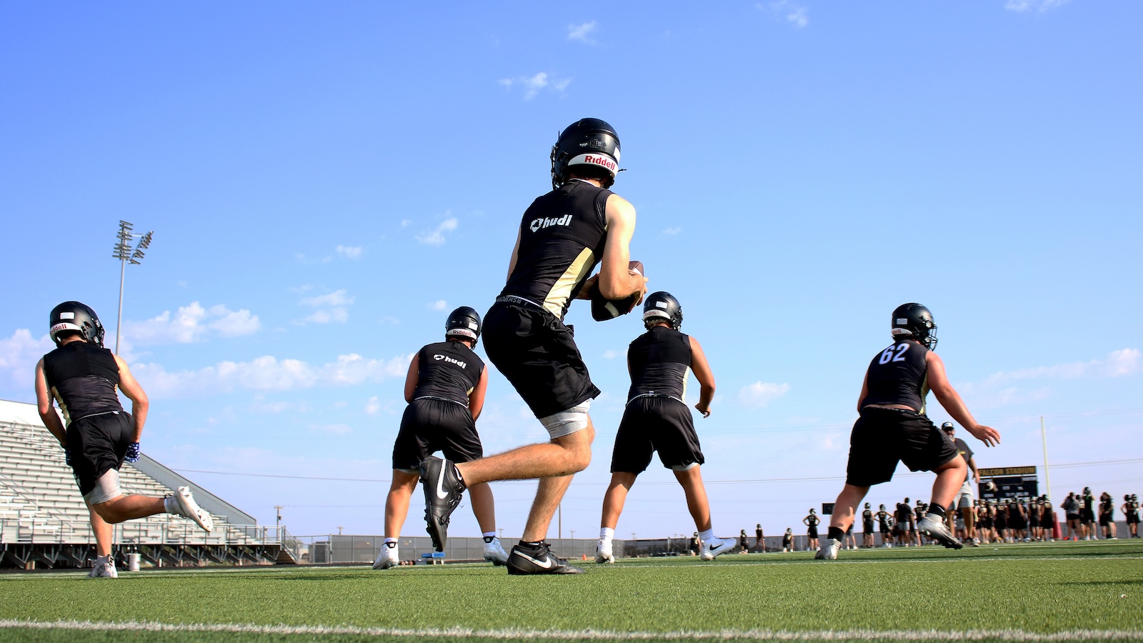 American football players in black uniforms practice on a sunny field; central player leaps with the ball as teammates position around him.