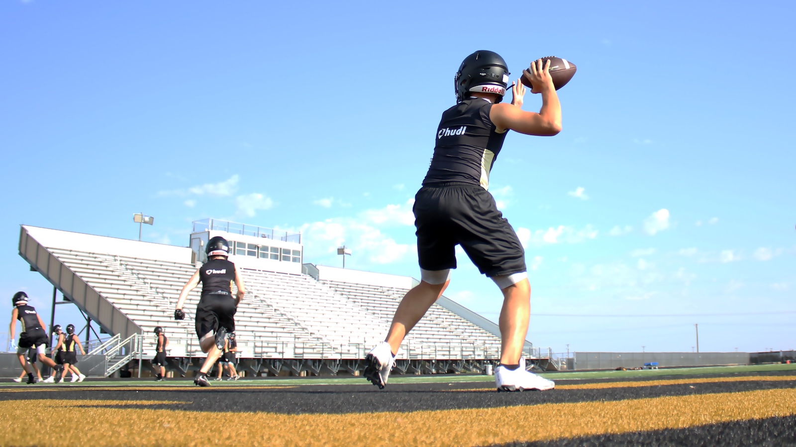 Quarterback in black uniform throws a football on a high school track field with empty bleachers in the background.