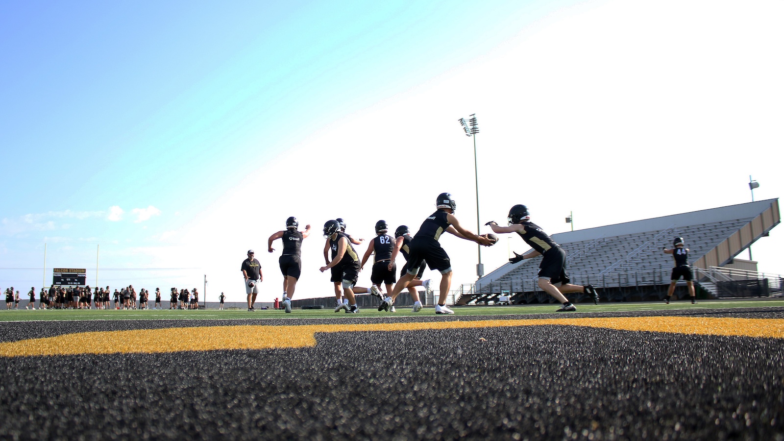 Football players in black uniforms practice on a turf field; a handoff occurs near the line of scrimmage with empty bleachers in the background under a clear sky.