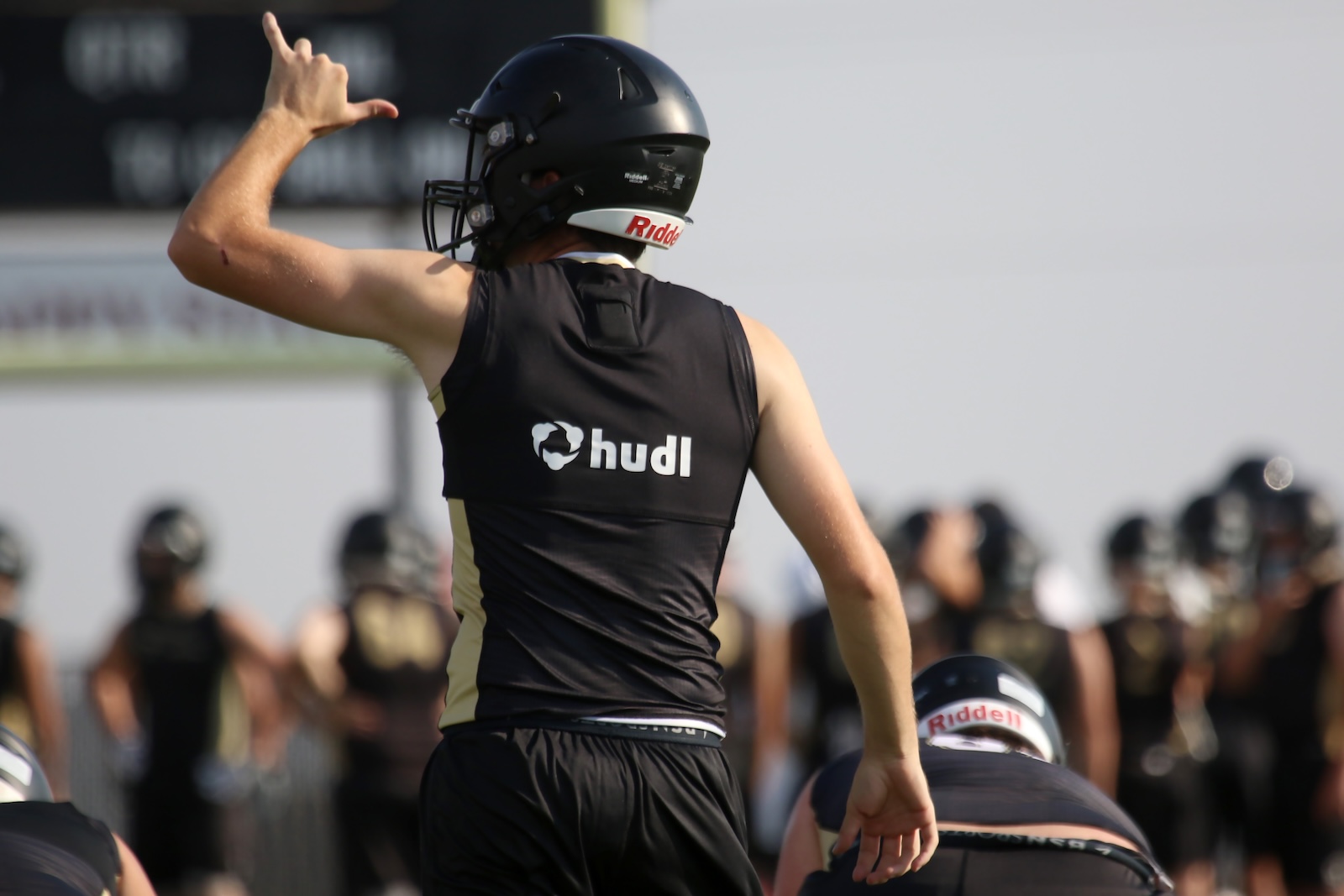 Football player in a black Hudl jersey giving a thumbs-up with helmet on during practice; teammates blurred in the background.