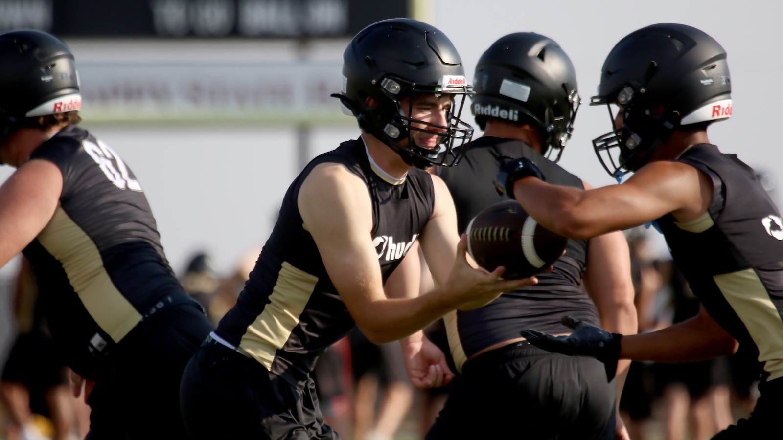 Football players in black and gold uniforms practice a drill as one player hands off a football to a teammate during a drill on a sunny field.