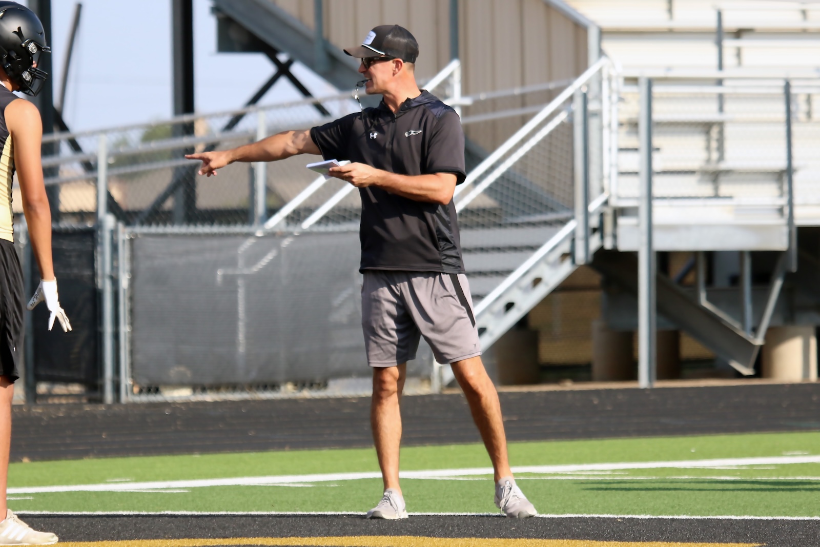 Coach in a black polo and cap points and holds play sheets as he directs players on a football field, with a helmeted teammate nearby.