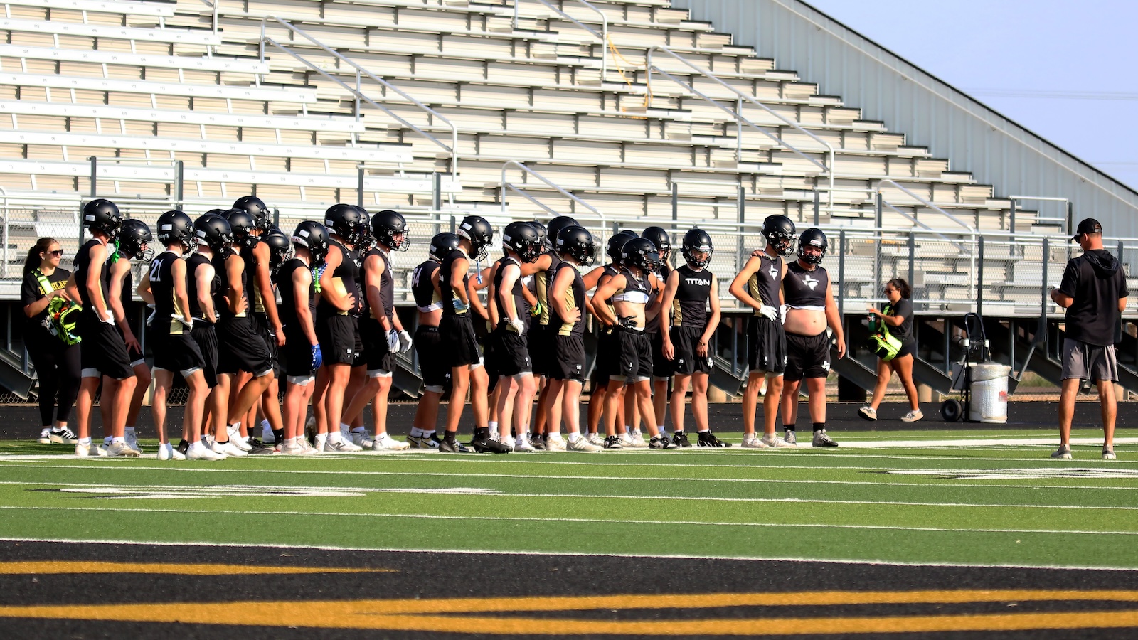 High school football team lined up in black uniforms and helmets on a football field, with a coach nearby.