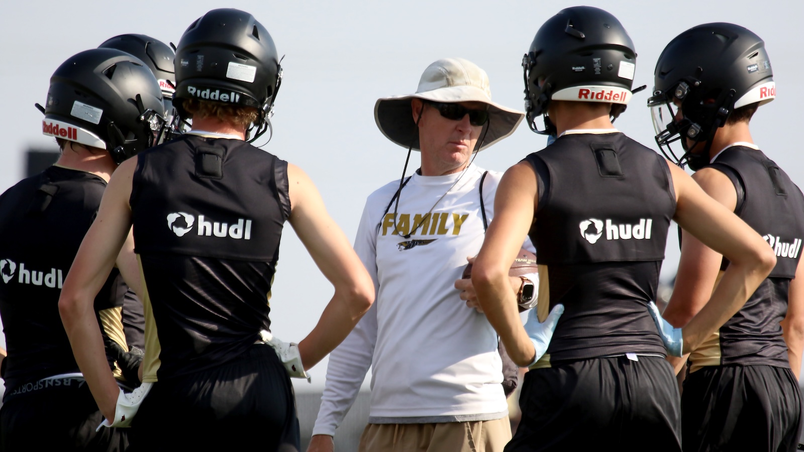 Football coach wearing a wide-brim hat and sunglasses talks to a circle of players in black Hudl vests during practice. The shirt reads ‘FAMILY’.