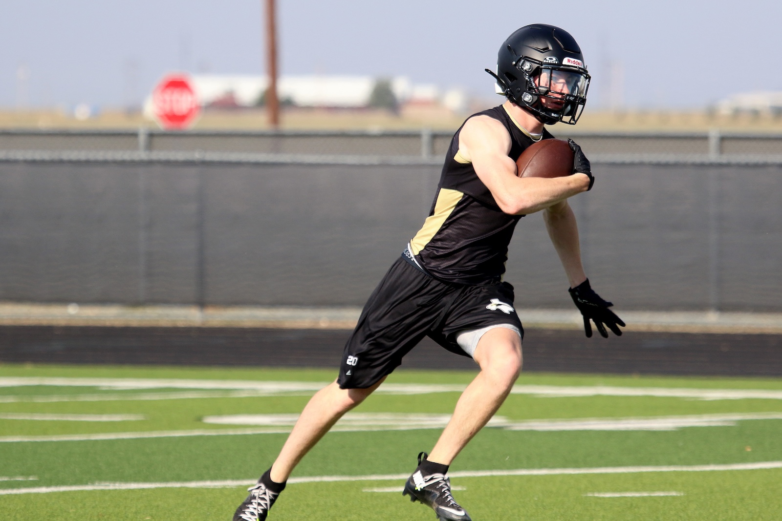 American football player in black and gold uniform running with the ball on a turf field, wearing a black helmet and gloves