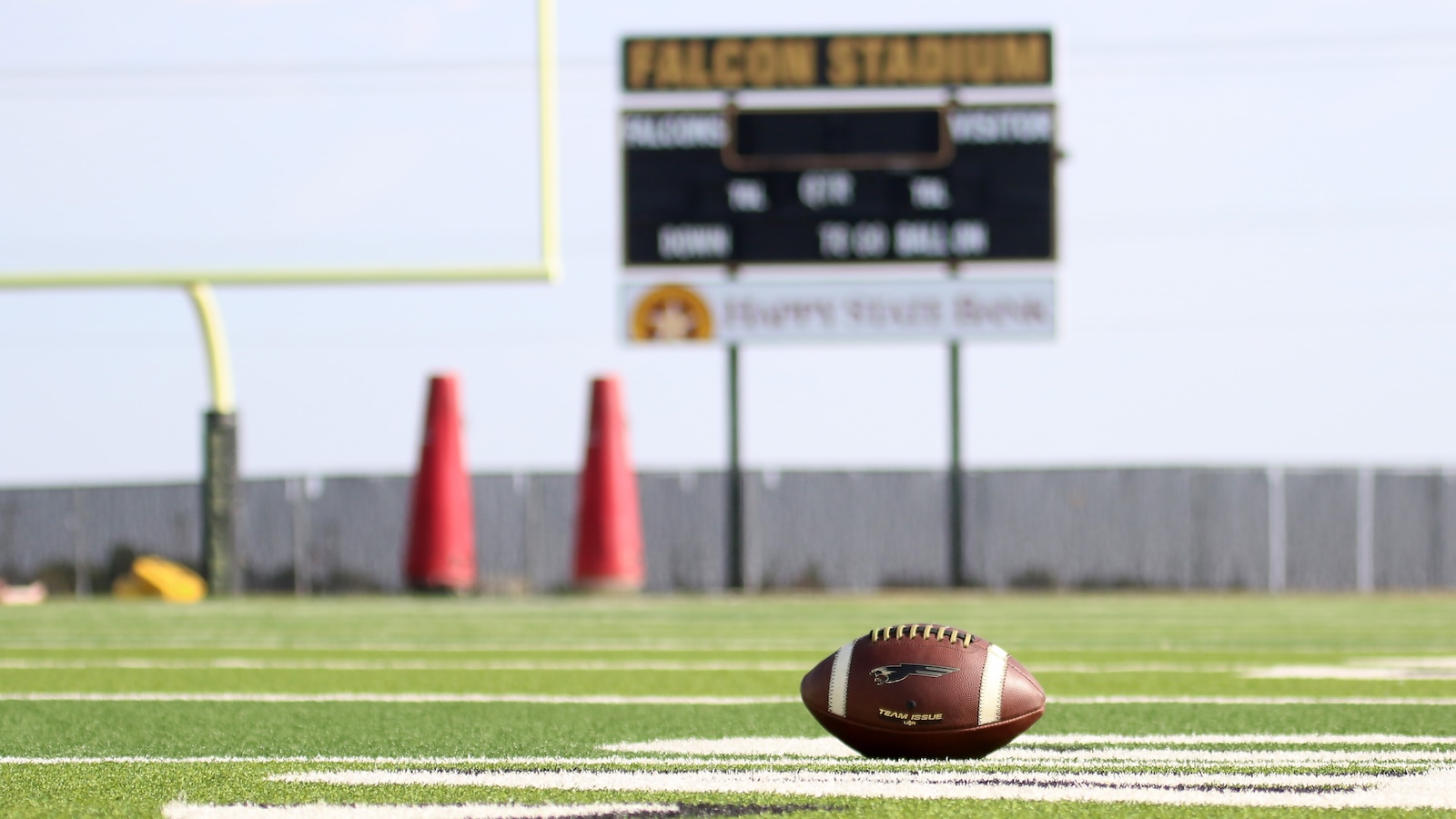 American football resting on a green turf field with a blurred scoreboard and goal posts in the background in daylight.