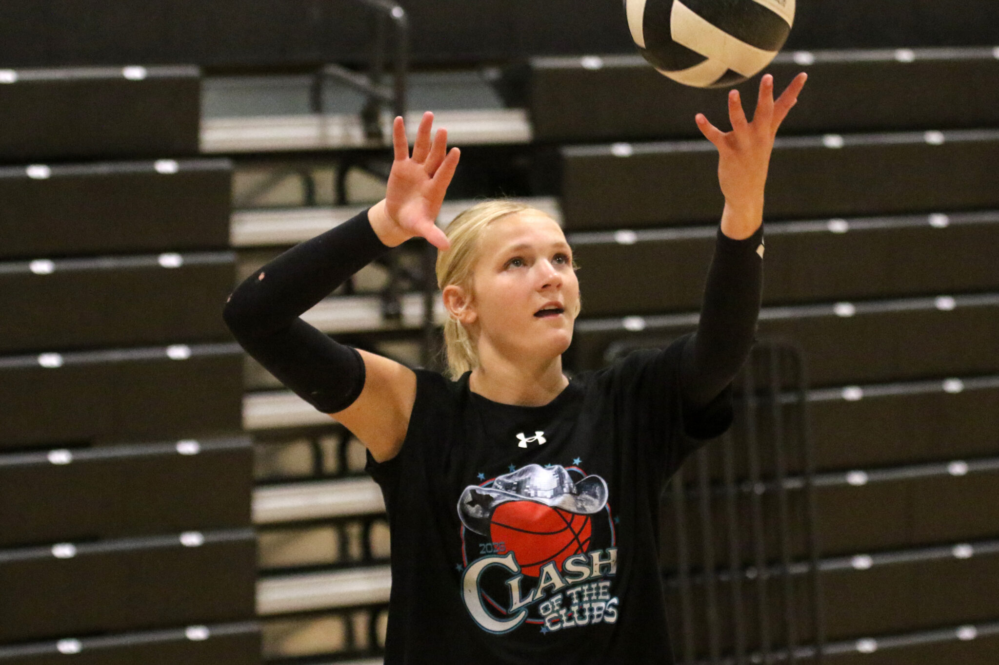 1K6A6012 | Press Pass Sports Female volleyball player in a black long-sleeve shirt reaching up to set a ball in a gym with bleachers in the background.