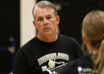 Middle-aged man with gray hair in a black Bushland volleyball t-shirt, looking to the left in a gym setting.
