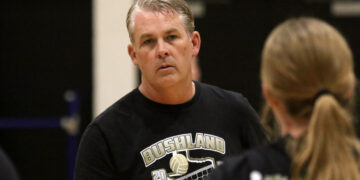 Middle-aged man with gray hair in a black Bushland volleyball t-shirt, looking to the left in a gym setting.