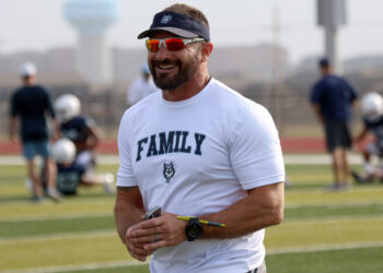 West Plains - Jeff Lofton 1 | Press Pass Sports Smiling man in a white 'FAMILY' shirt and visor stands on a football field during practice, holding a few writing implements and wearing orange sunglasses and a wristwatch.