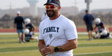 Smiling man in a white 'FAMILY' shirt and visor stands on a football field during practice, holding a few writing implements and wearing orange sunglasses and a wristwatch.
