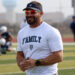 West Plains - Jeff Lofton 1 | Press Pass Sports Smiling man in a white 'FAMILY' shirt and visor stands on a football field during practice, holding a few writing implements and wearing orange sunglasses and a wristwatch.
