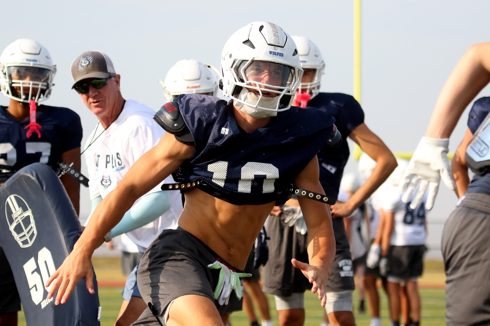 Football practice scene with a central player in navy jersey #10 and white helmet lunging forward on a field, coach and teammates behind him.