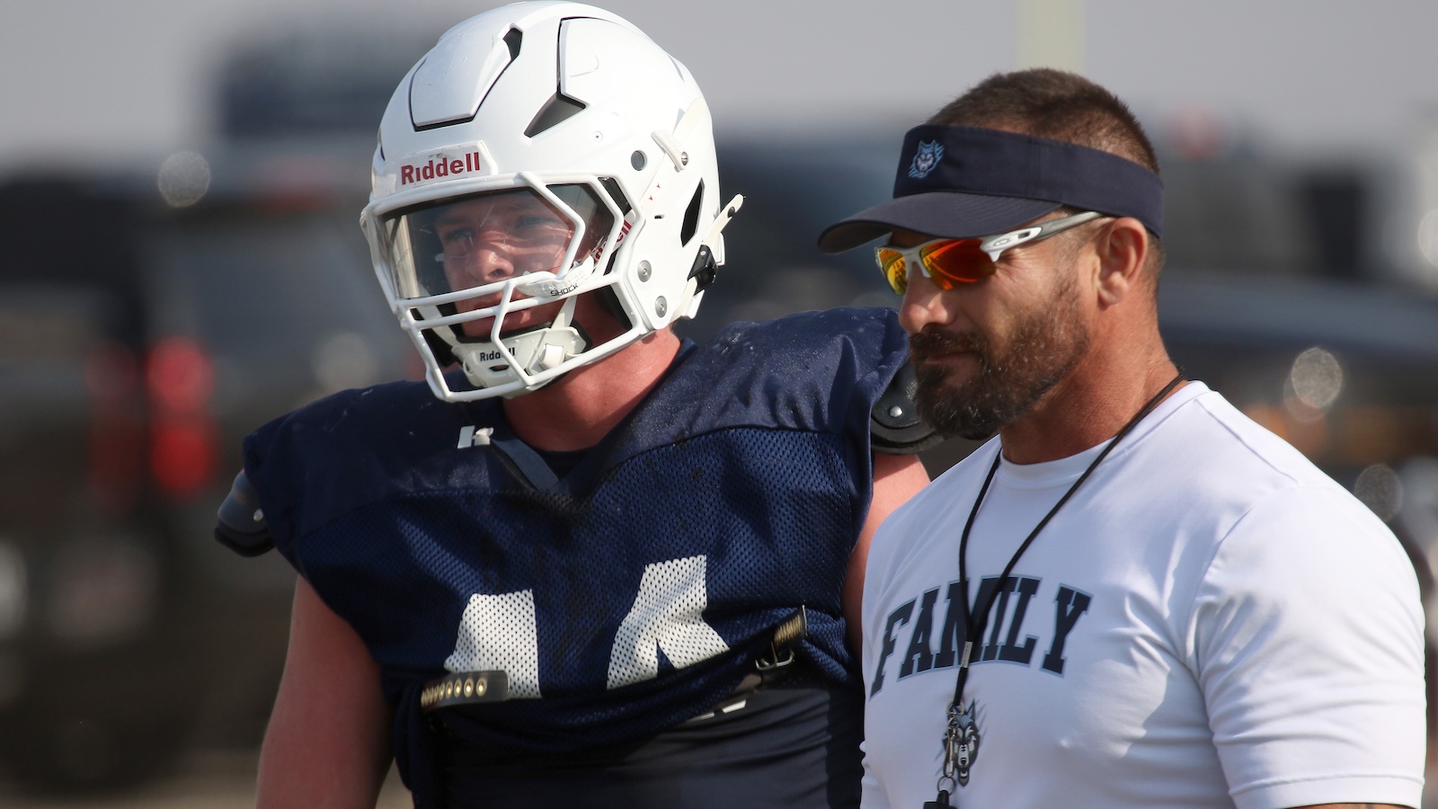 Football player in navy practice gear with a white helmet standing beside a bearded coach in a white shirt outdoors.