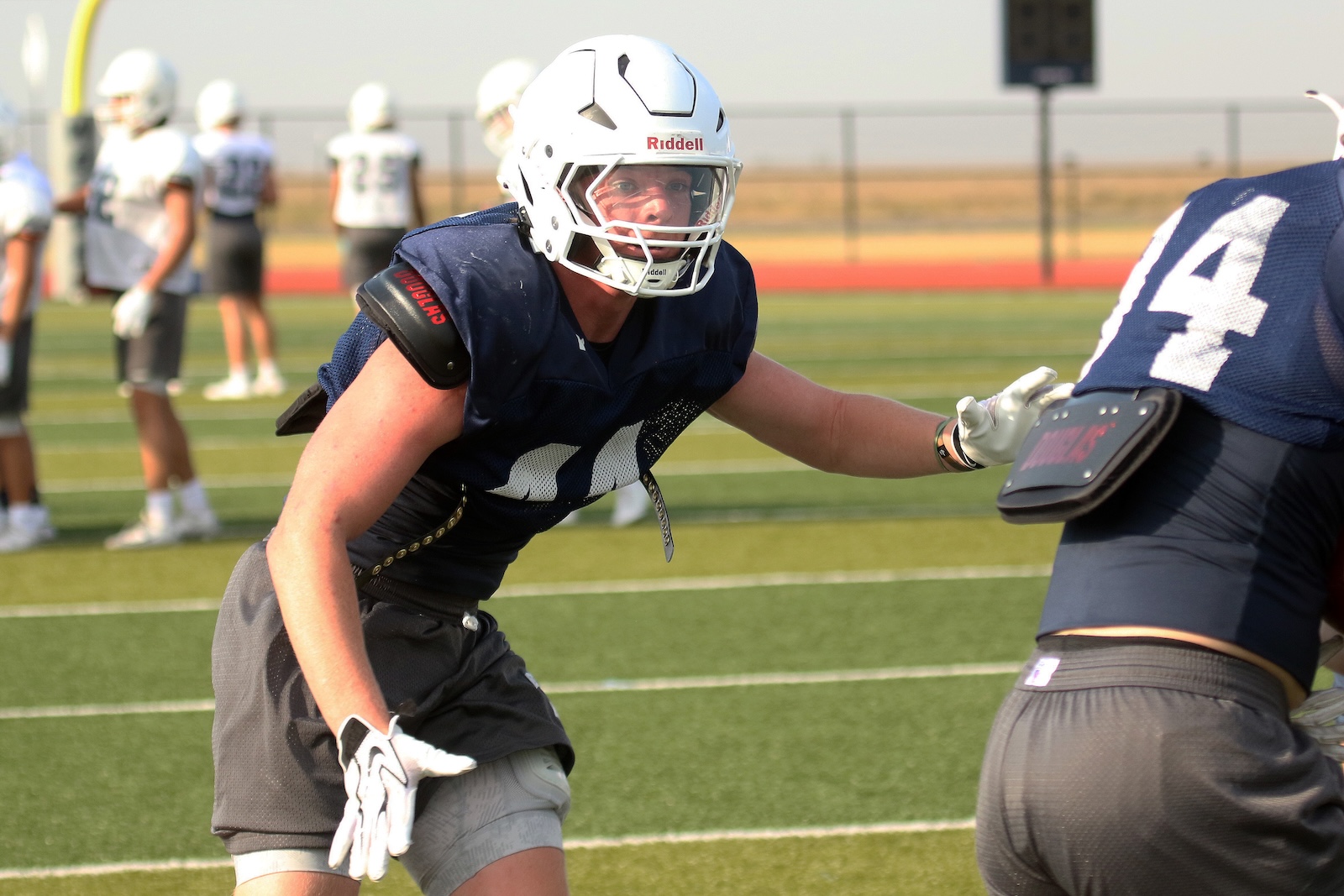Football practice scene: player in navy jersey and white helmet lunges forward with outstretched arm on artificial turf field.