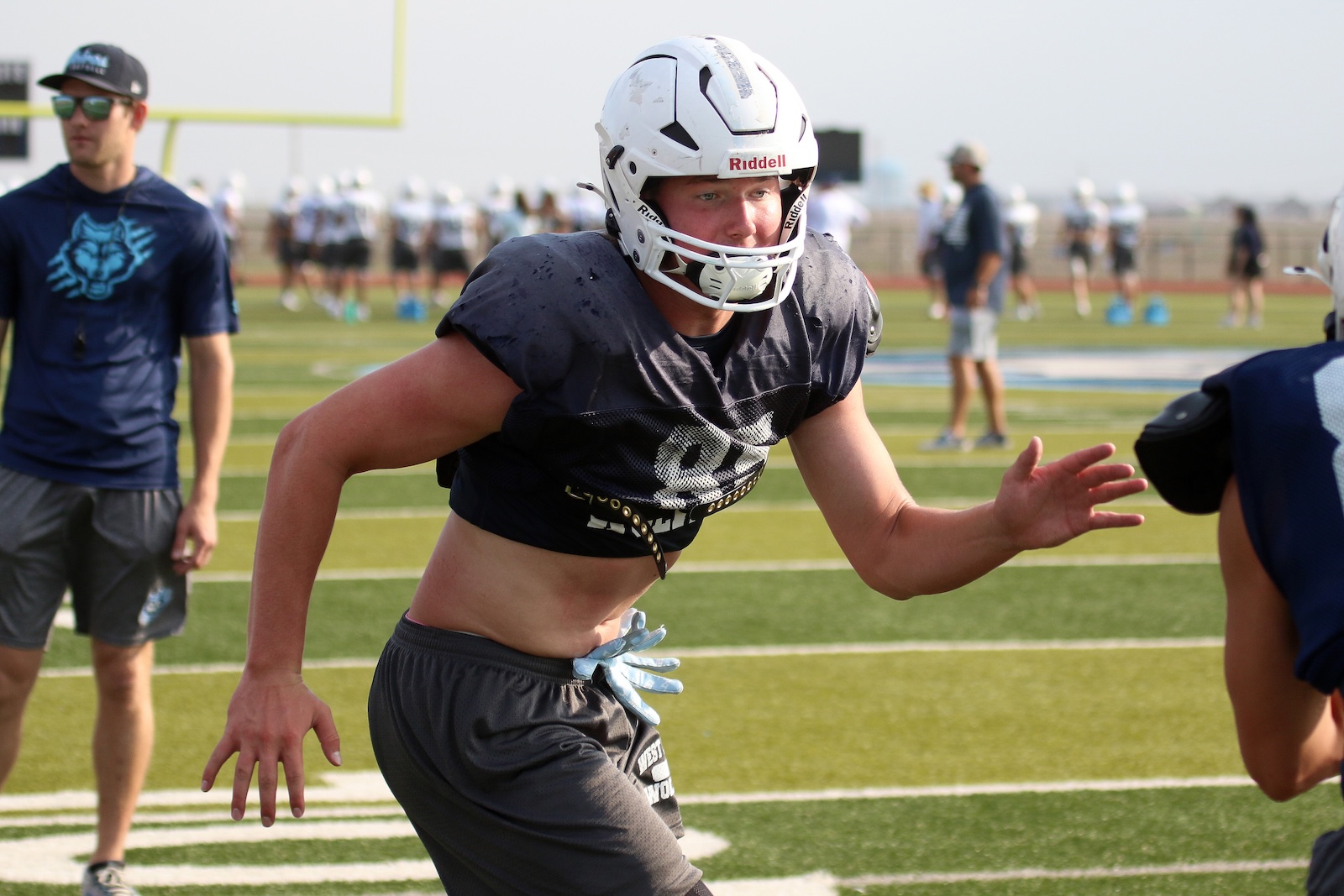 Football player in a white helmet and black practice jersey sprinting on a synthetic turf field during drills, teammates in the background.