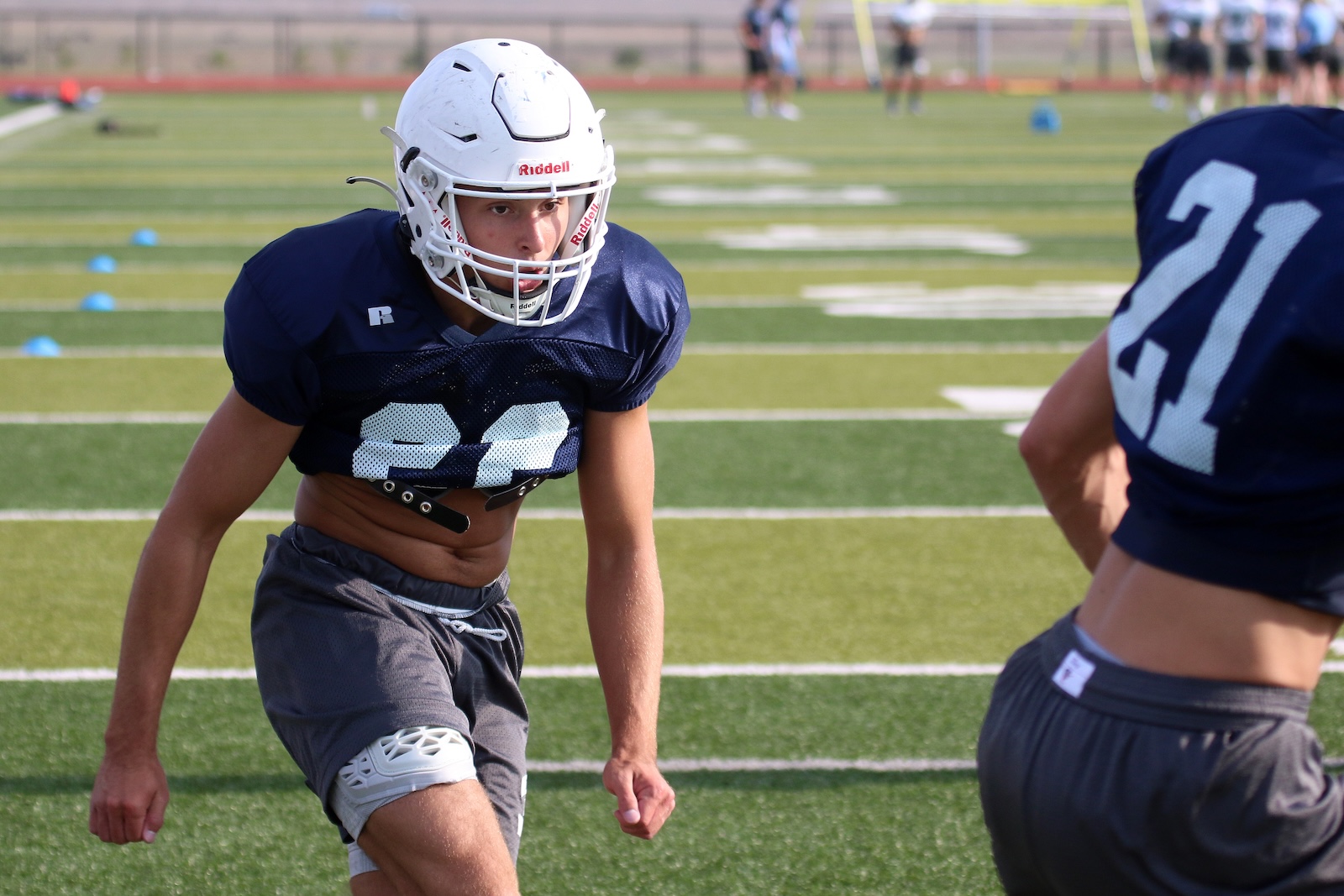Football practice scene: a player in navy jersey #22 wearing a white helmet, crouched and ready on a green turf field.