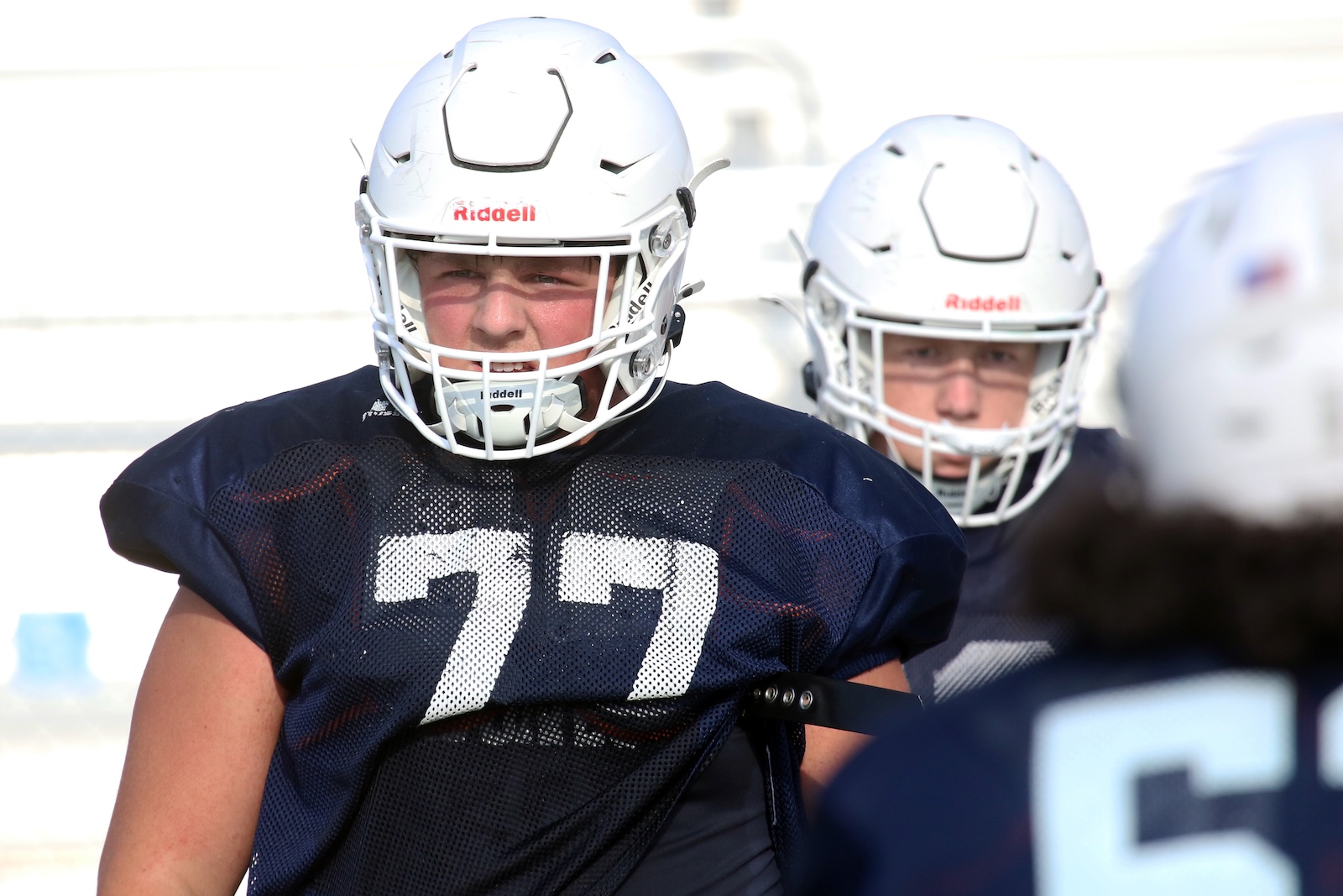Two football players in navy practice jerseys and white helmets on a sunny field, facing the camera in a drill.
