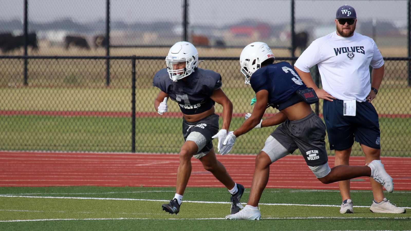 Two football players in navy uniforms sprint toward the left on a turf field as a coach watches from the right, wearing a white Wolves shirt and cap.