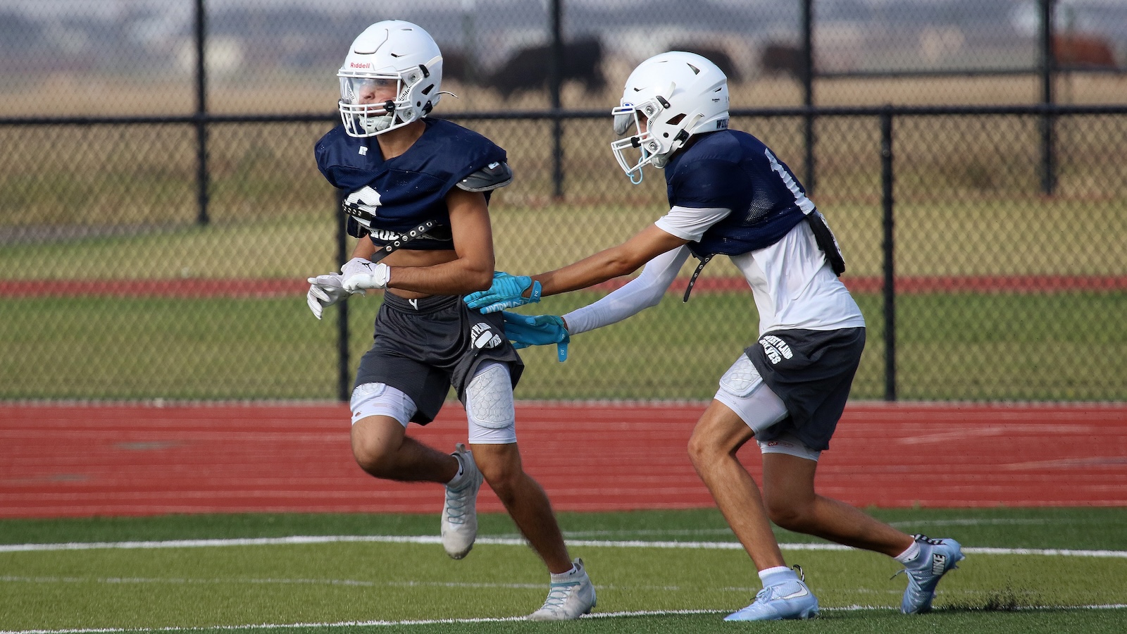 Two high school football players in navy uniforms run on a football field, with a chain-link fence in the background, during practice or a game.