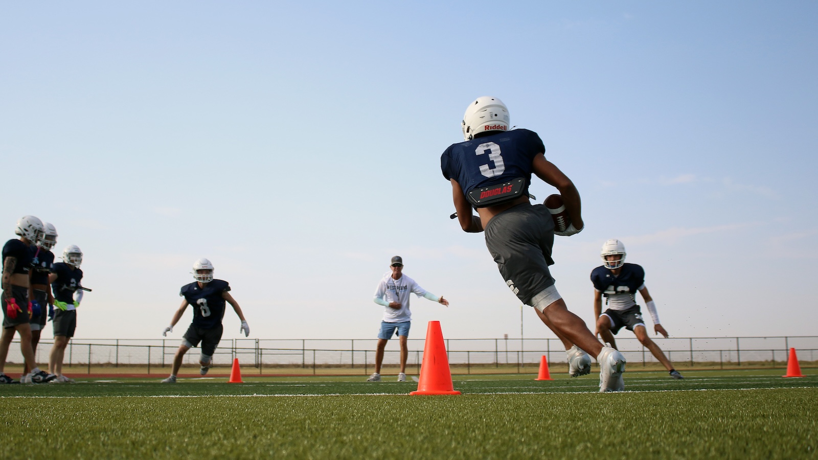 Young football players in helmets practice drills on a field, one leaping with the ball near orange cones